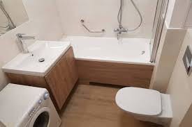 Jasna łazienka po remoncie: umywalka z szafką w kolorze drewna, biała wanna z parawanem nawannowym, toaleta i pralka. Widoczne relingi dla osób z ograniczoną sprawnością ruchową.