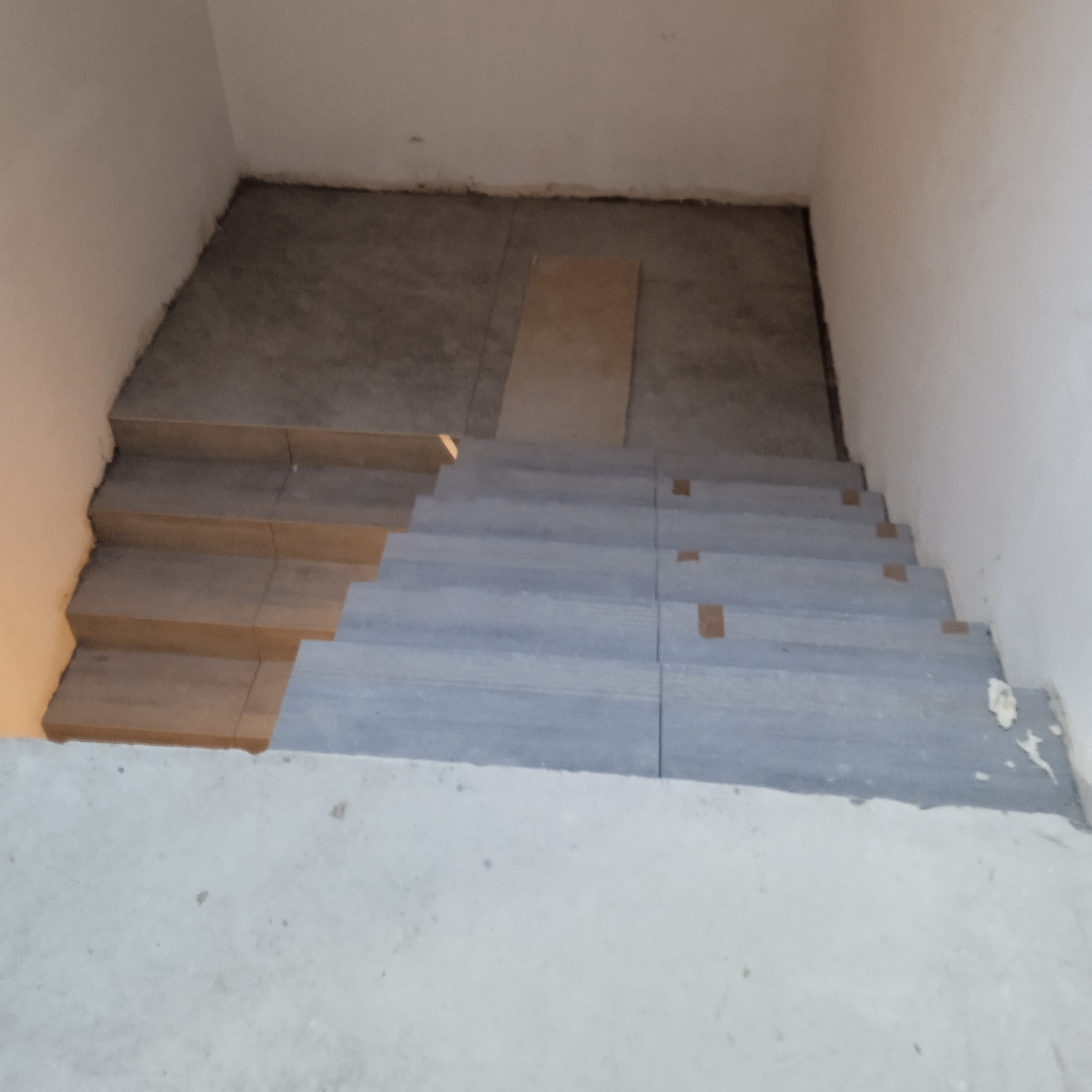 Wnętrze w trakcie prac wykończeniowych: częściowo wyłożone płytkami schody prowadzące na wyłożony płytkami podest, widoczne fragmenty taśmy malarskiej i niezakończone fugowanie.