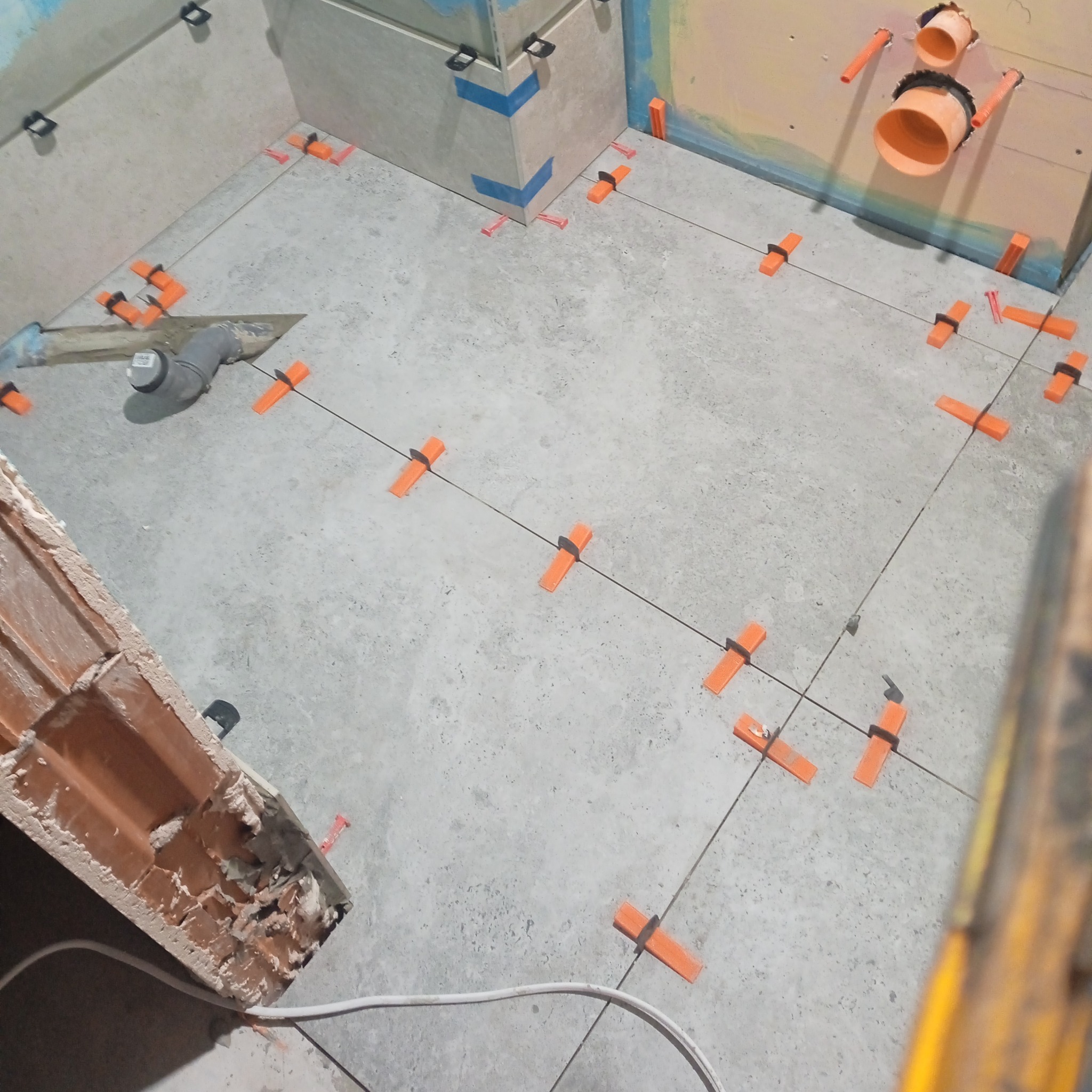 Układanie płytek podłogowych z użyciem pomarańczowych klipsów poziomujących, widoczne rury kanalizacyjne w ścianie oraz fragment cegły i kielni