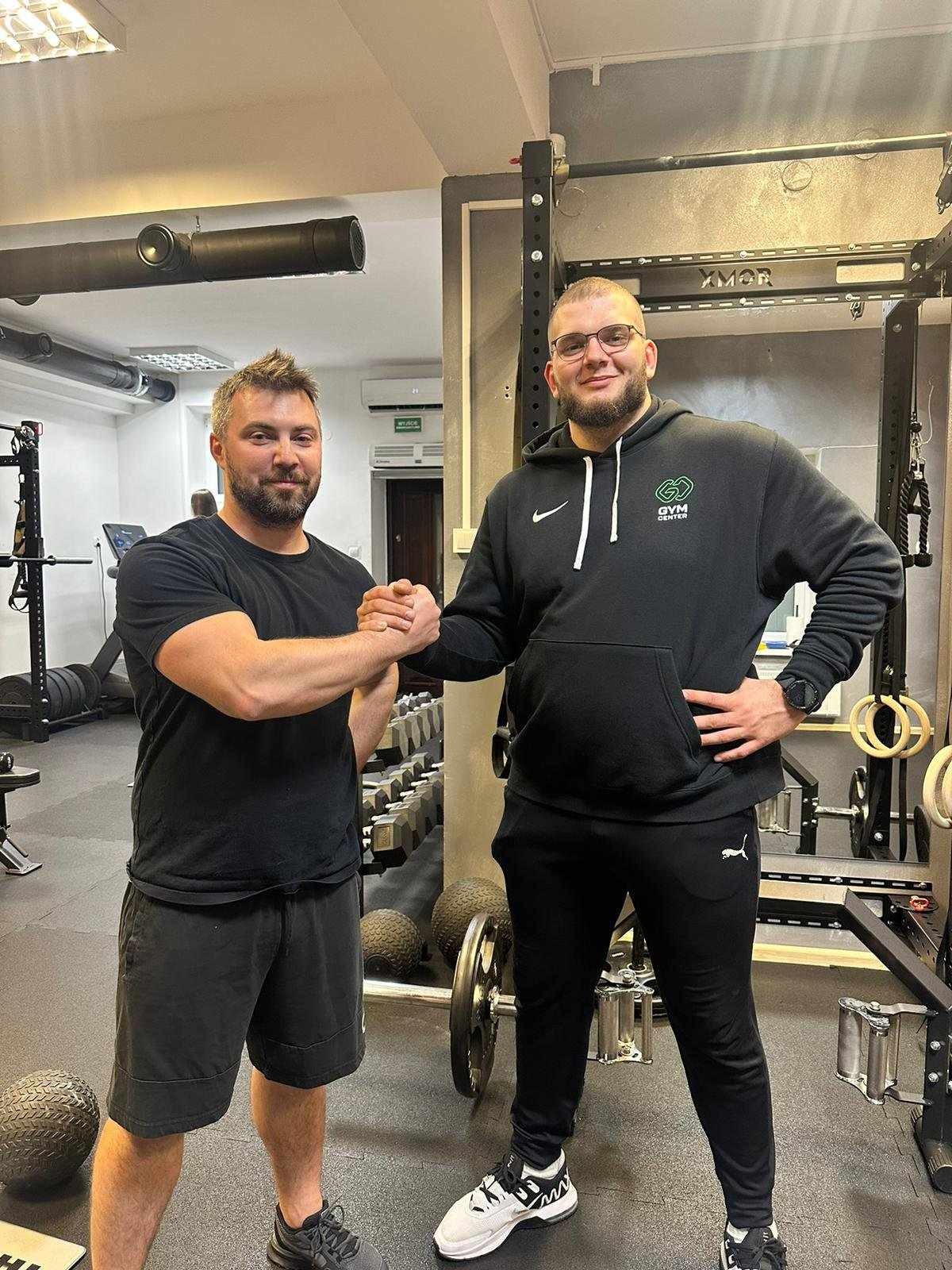 Dwóch mężczyzn w sportowych strojach ściska sobie dłonie na tle siłowni z widocznym sprzętem do ćwiczeń siłowych i odważnikami.