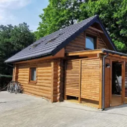 Drewniany dom z bali z dobudowaną altaną, szary dach z widocznymi oknami dachowymi, kostka brukowa, rowery opierające się o ścianę, otoczenie zieleni.