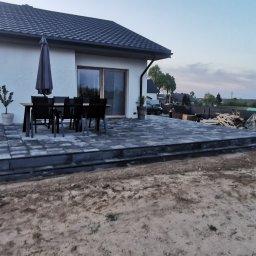 Perfect Haus - Nowoczesny dom z tarasem wyłożonym szarą kostką brukową. Widoczne meble ogrodowe, parasol, trawnik w trakcie zakładania. W tle inne budynki i zieleń.