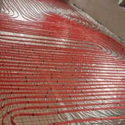 Czerwone rury ogrzewania podłogowego ułożone na folii w surowym wnętrzu. Widoczne okna i ściany w trakcie wykończenia. Instalacja przed wylaniem posadzki.