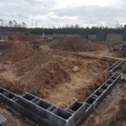 Wykop pod fundamenty z ułożonymi bloczkami betonowymi. Widoczny wykop w kształcie litery L, stos bloczków i biały bus w tle, pochmurne niebo.