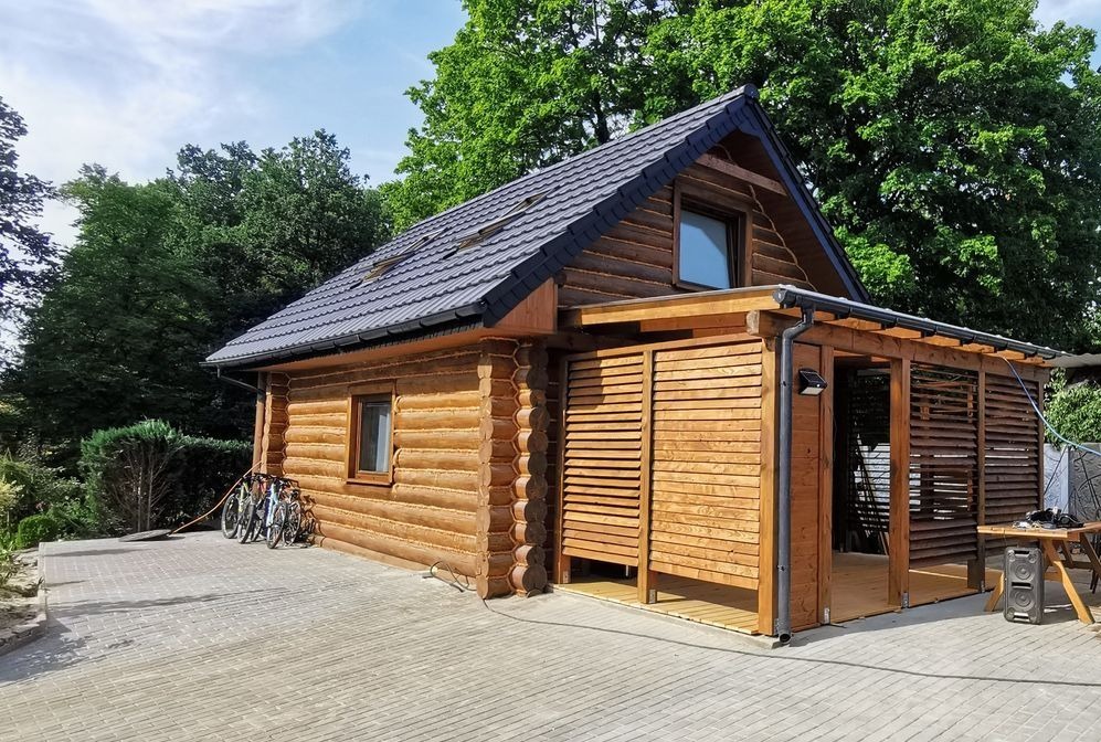 Drewniany dom z bali z dobudowaną altaną, szary dach z widocznymi oknami dachowymi, kostka brukowa, rowery opierające się o ścianę, otoczenie zieleni.