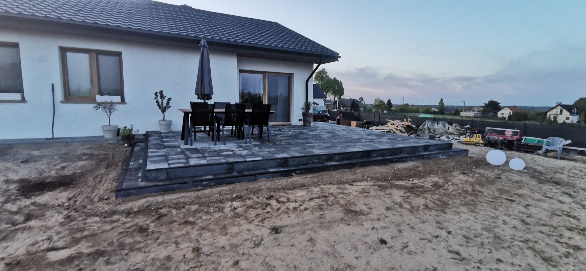 Nowoczesny dom z tarasem wyłożonym szarą kostką brukową. Widoczne meble ogrodowe, parasol, trawnik w trakcie zakładania. W tle inne budynki i zieleń.