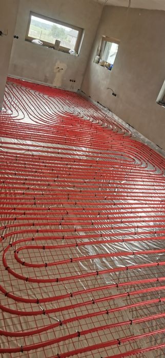 Czerwone rury ogrzewania podłogowego ułożone na folii w surowym wnętrzu. Widoczne okna i ściany w trakcie wykończenia. Instalacja przed wylaniem posadzki.