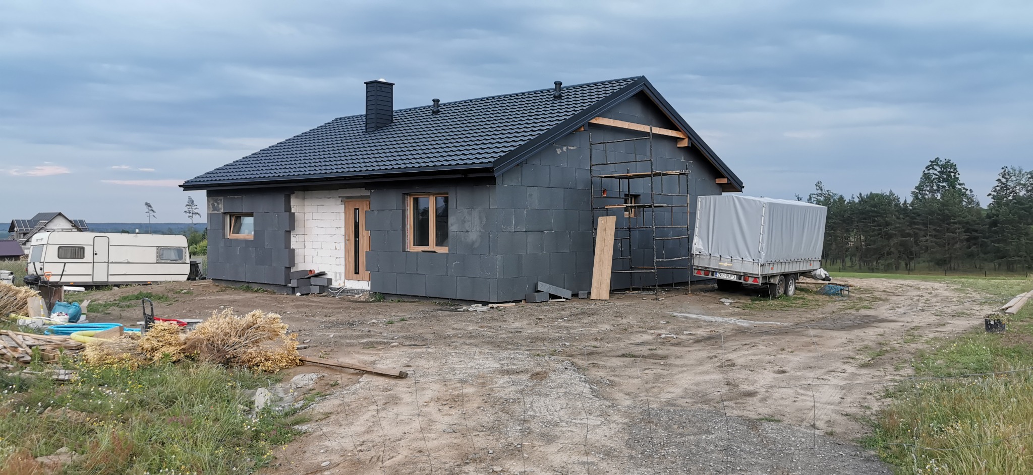 Niedokończony dom z szarych bloczków, z czarnym dachem, widoczne rusztowanie i przyczepa. Część ścian jest ocieplona białym materiałem. Na pierwszym planie ziemia.