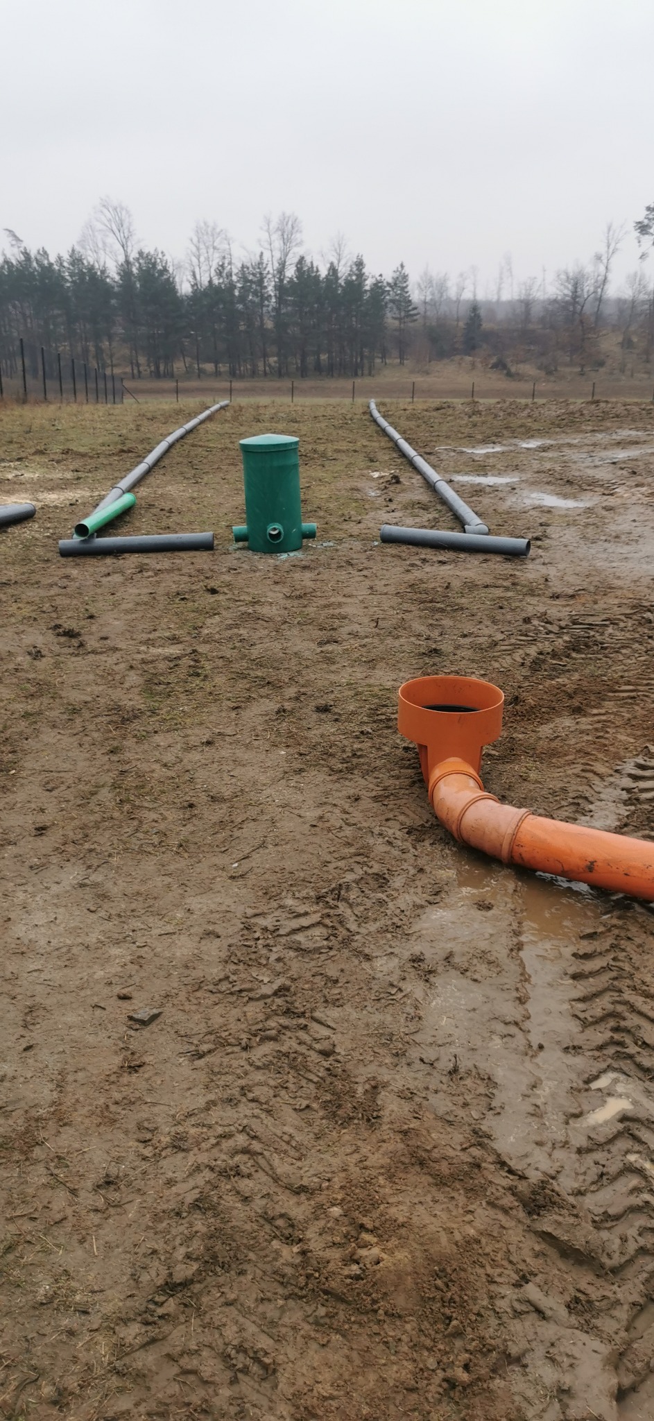 Instalacja kanalizacyjna z rur PVC na zewnątrz, widoczne elementy systemu w kolorach szarym, zielonym i pomarańczowym, ułożone na błotnistym terenie.