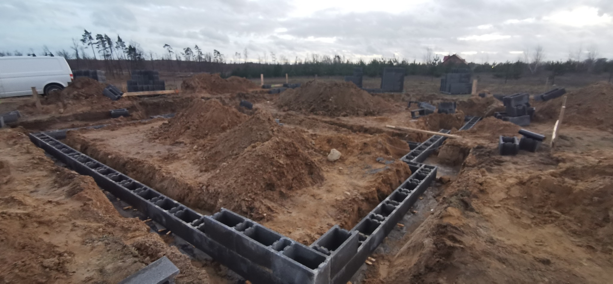 Wykop pod fundamenty z ułożonymi bloczkami betonowymi. Widoczny wykop w kształcie litery L, stos bloczków i biały bus w tle, pochmurne niebo.