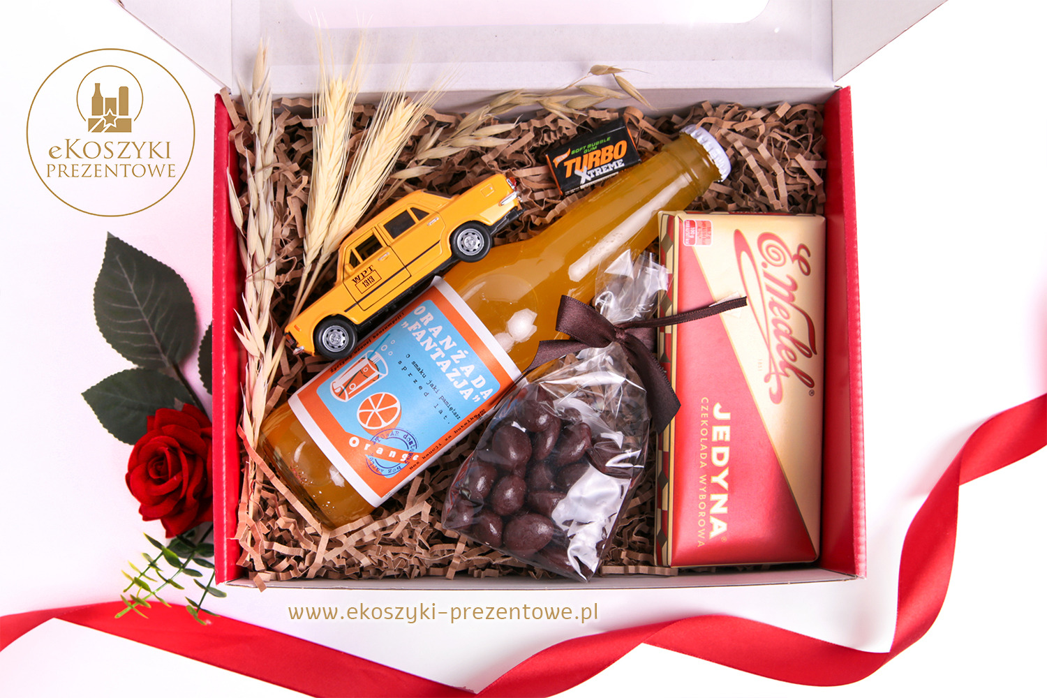 Otwarty, czerwono-biały box prezentowy z zawartością: oranżada, czekolada, czekoladki, zabawkowy taksówkarz i zboża. Całość przewiązana czerwoną wstążką.