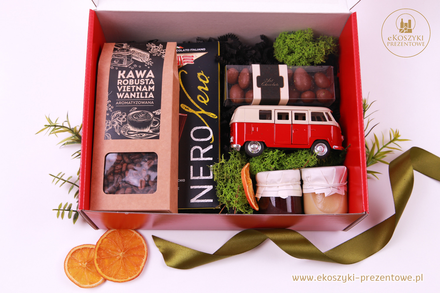 Czerwony kosz prezentowy z kawą, czekoladą, miodem, dekoracjami i modelem czerwonego VW busa na białym tle, ozdobiony zieloną wstążką i suszonymi pomarańczami.
