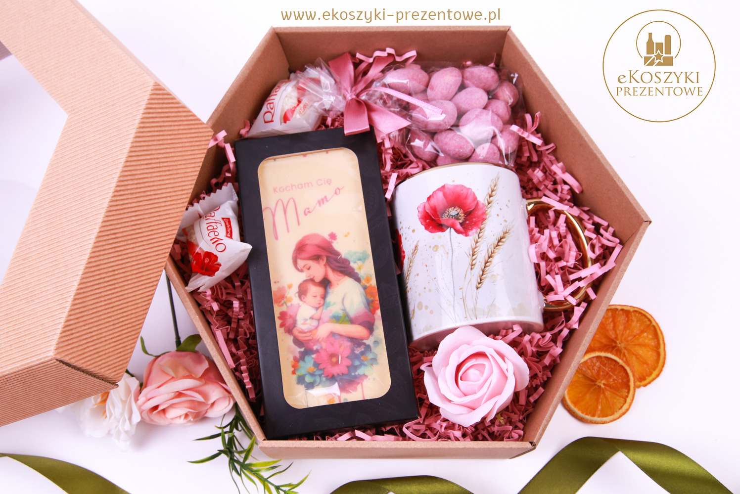 Otwarty, heksagonalny box prezentowy z czekoladą 'Kocham Cię Mamo', kubkiem z makiem, słodyczami i suszonymi pomarańczami, wypełniony różowym siankiem.