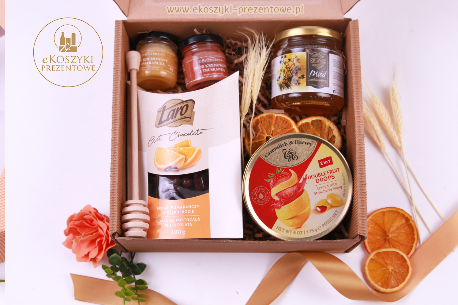 Elegancki kosz prezentowy z miodami, czekoladą, suszonymi pomarańczami i cukierkami, ozdobiony kwiatem i wstążką, idealny na upominek.