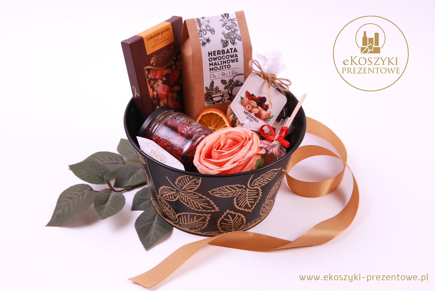 Elegancki, czarny kosz z motywem liści wypełniony słodkościami: czekoladą, herbatą malinową, miodem malinowym, mieszanką bakalii i lizakiem, ozdobiony różą i wstążką.
