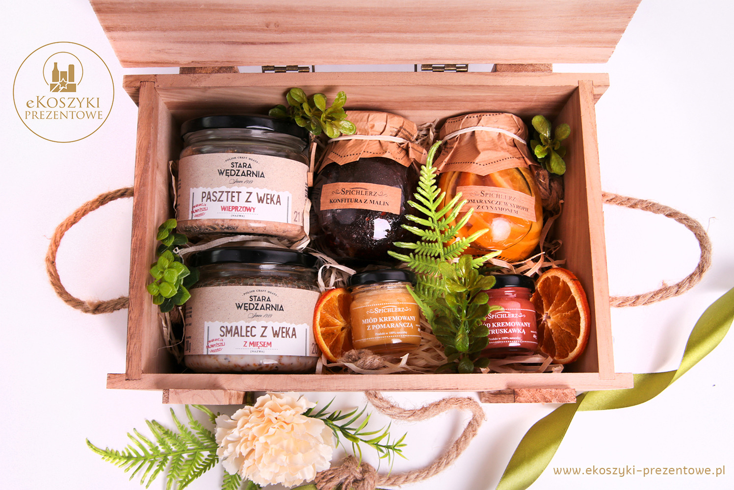 Drewniana skrzynka z różnorodnymi przetworami: pasztet, smalec, konfitura, miód, udekorowana zielonymi gałązkami i suszonymi pomarańczami.