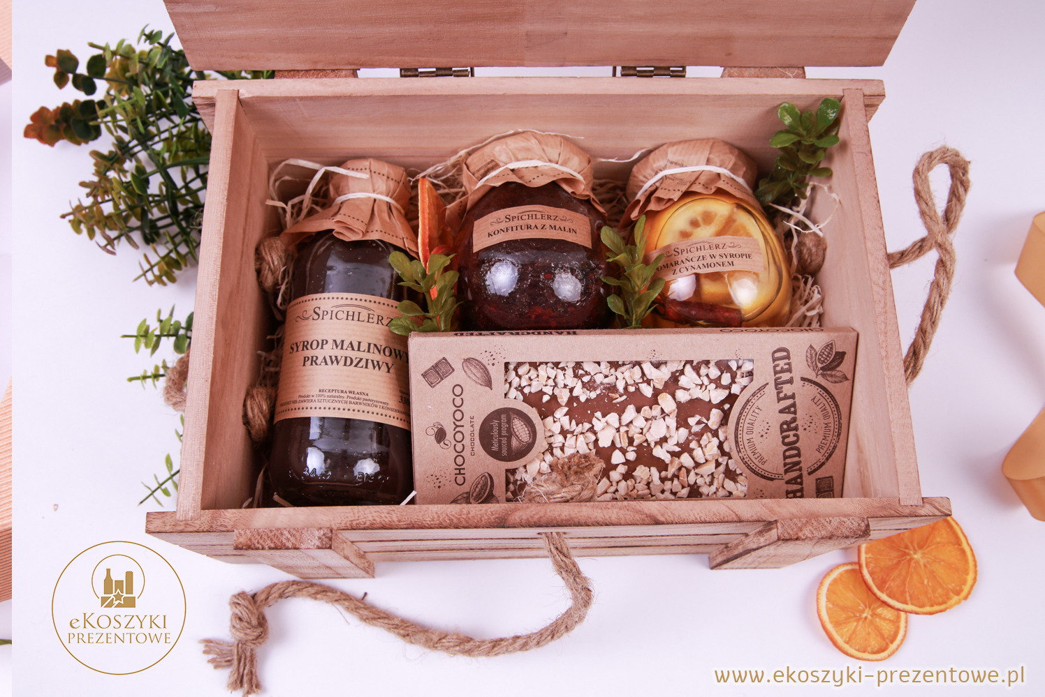 Otwarty drewniany kosz prezentowy z syropem malinowym, konfiturą z malin, pomarańczami w syropie, czekoladą i dekoracjami roślinnymi na białym tle.