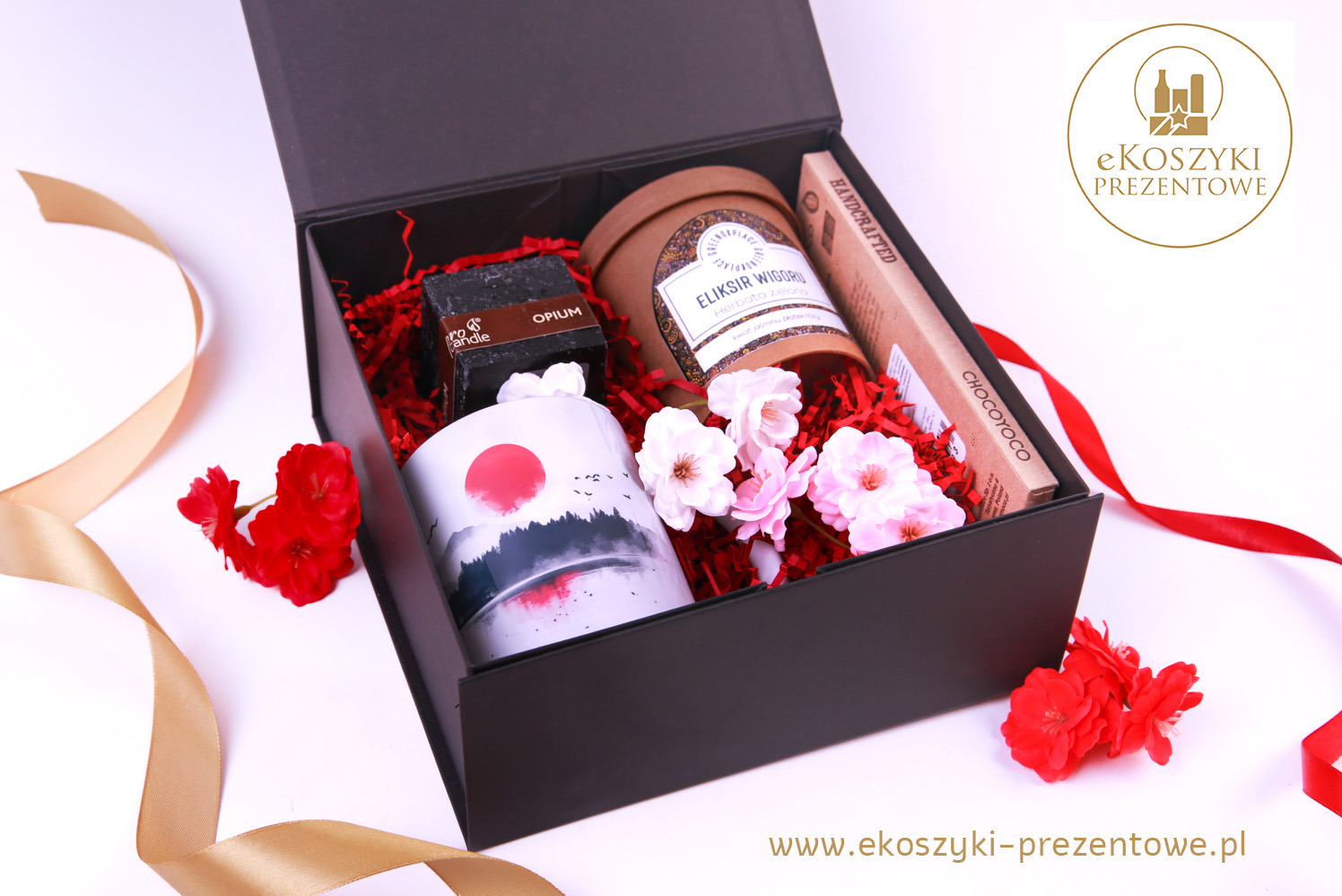 Czarny, otwarty box prezentowy wypełniony ozdobnym wypełniaczem, świecą, herbatą, mydłem i czekoladą, udekorowany kwiatami i wstążkami na białym tle.
