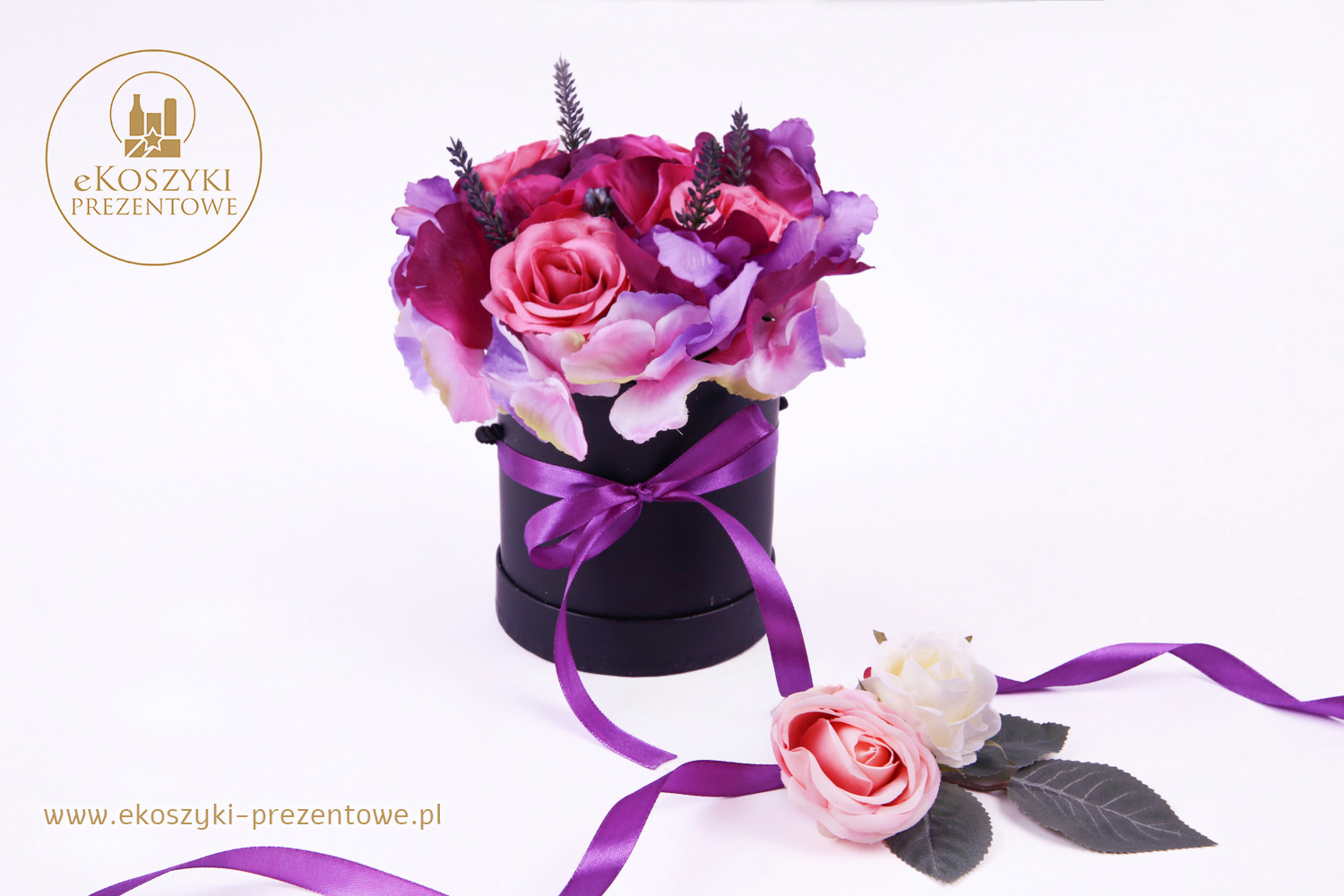 Kompozycja kwiatowa w czarnym pudełku, przewiązana fioletową wstążką, z dodatkowymi kwiatami i wstążką leżącymi obok na białym tle.