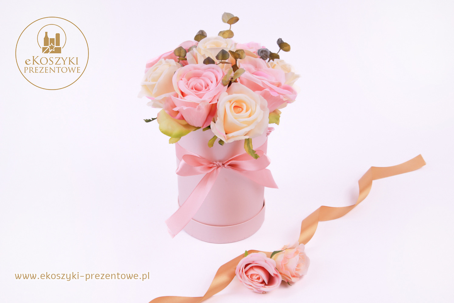 Różowe róże w okrągłym pudełku przewiązanym wstążką, leżąca złota wstążka i dwie róże na białym tle. Elegancka kompozycja kwiatowa.