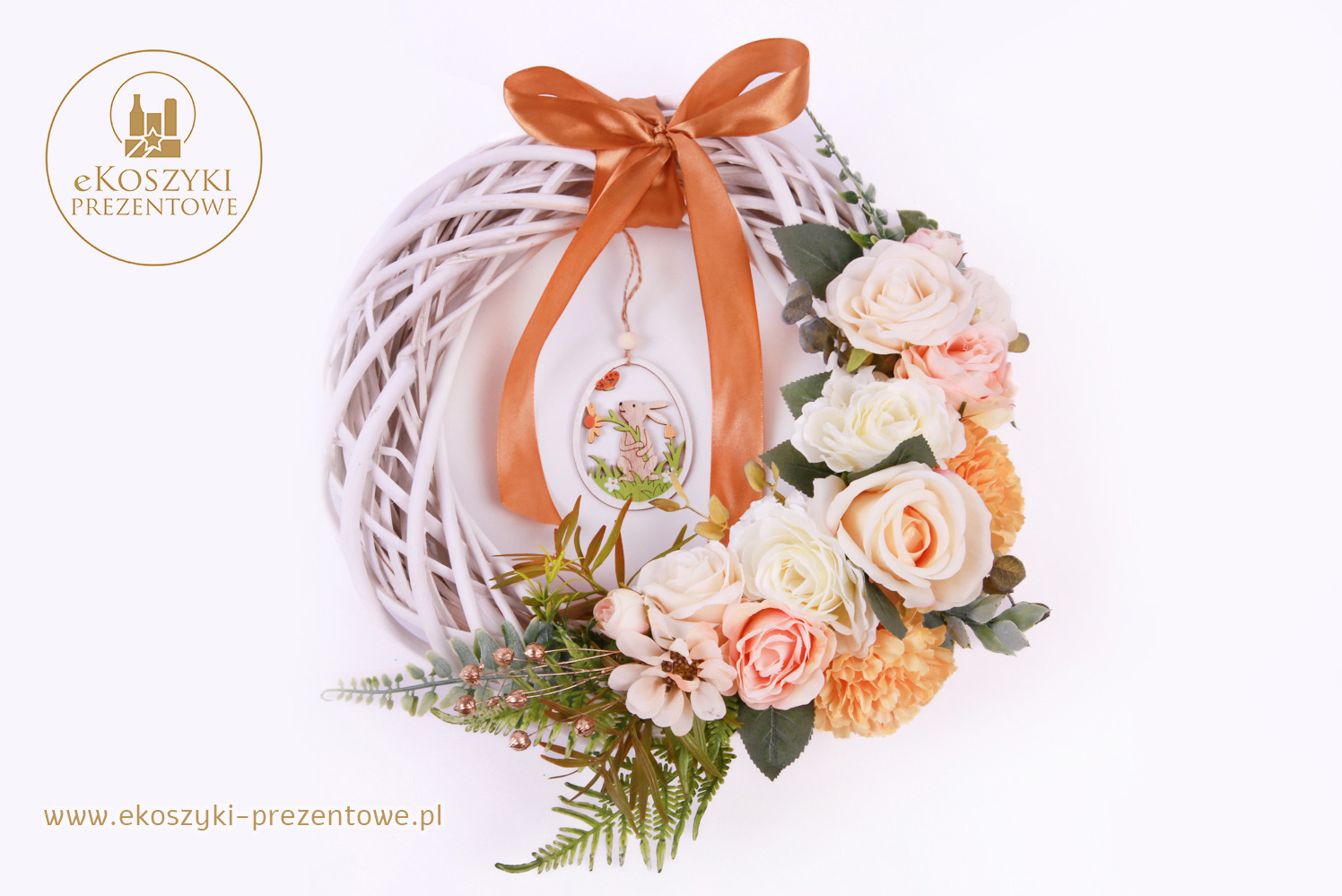 Ręcznie robiony biały wieniec z wikliny, zdobiony pastelowymi różami, zielonymi listkami i brązową wstążką. Zawieszka z zajączkiem dodaje uroku.
