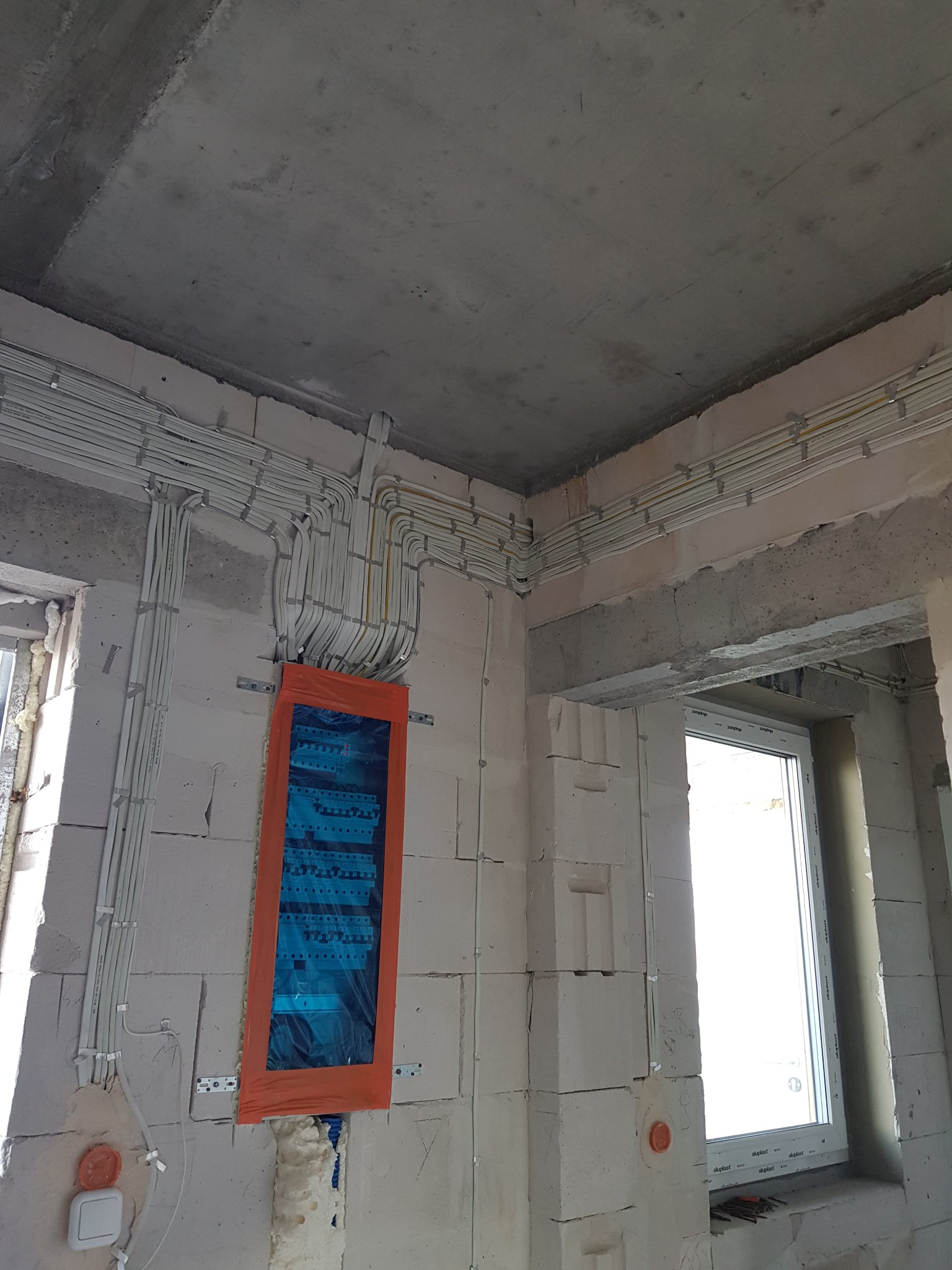 Surowe wnętrze w trakcie prac wykończeniowych: widoczne kable elektryczne ułożone wzdłuż ścian pod sufitem, zabezpieczona folią rozdzielnica elektryczna na ścianie z betonu komórkowego, okna...