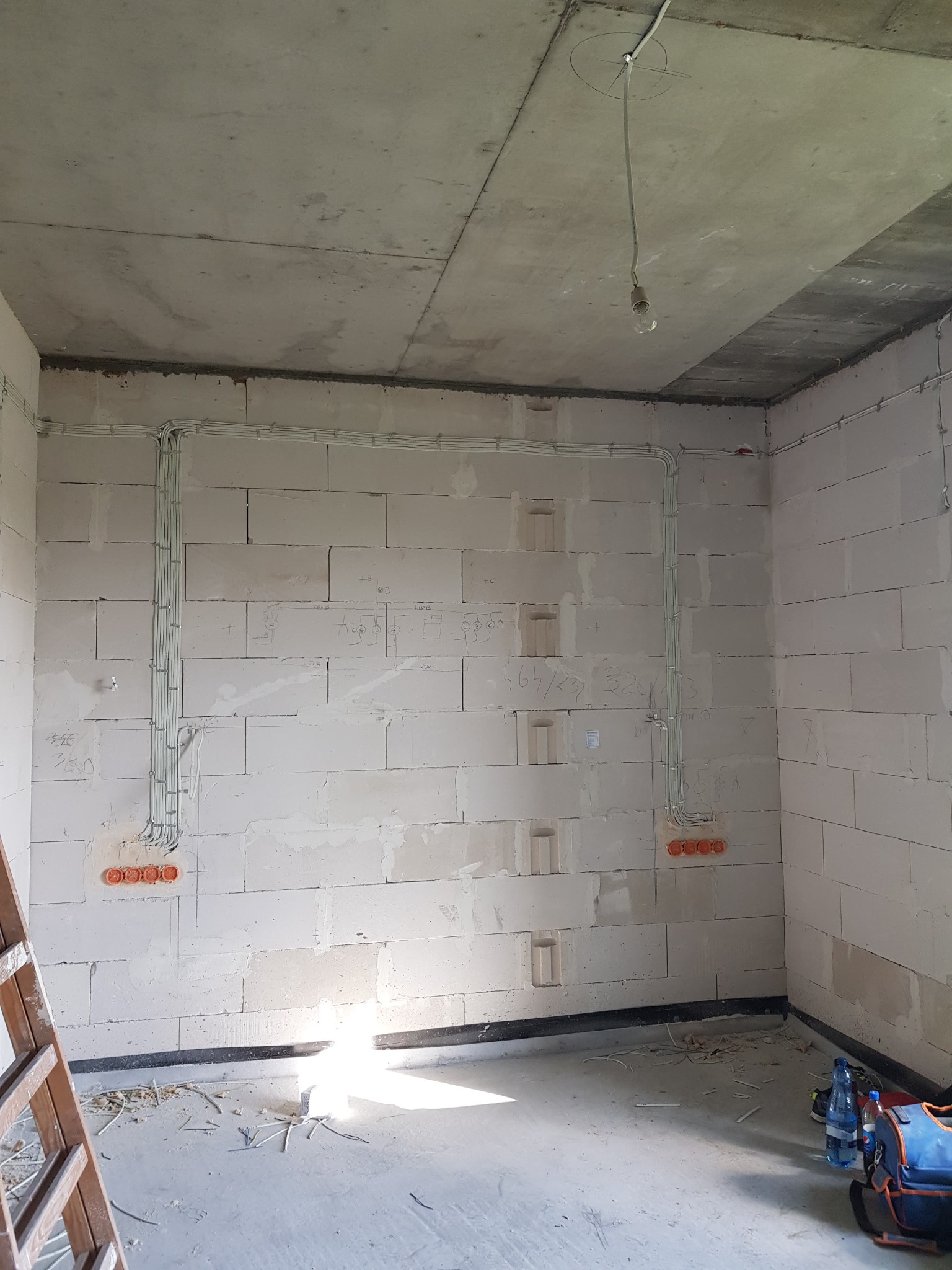 Surowe wnętrze w trakcie prac elektrycznych: widoczne kable prowadzone po ścianach z betonu komórkowego, puszki elektryczne w kolorze pomarańczowym, prowizoryczna żarówka zwisająca z sufitu...