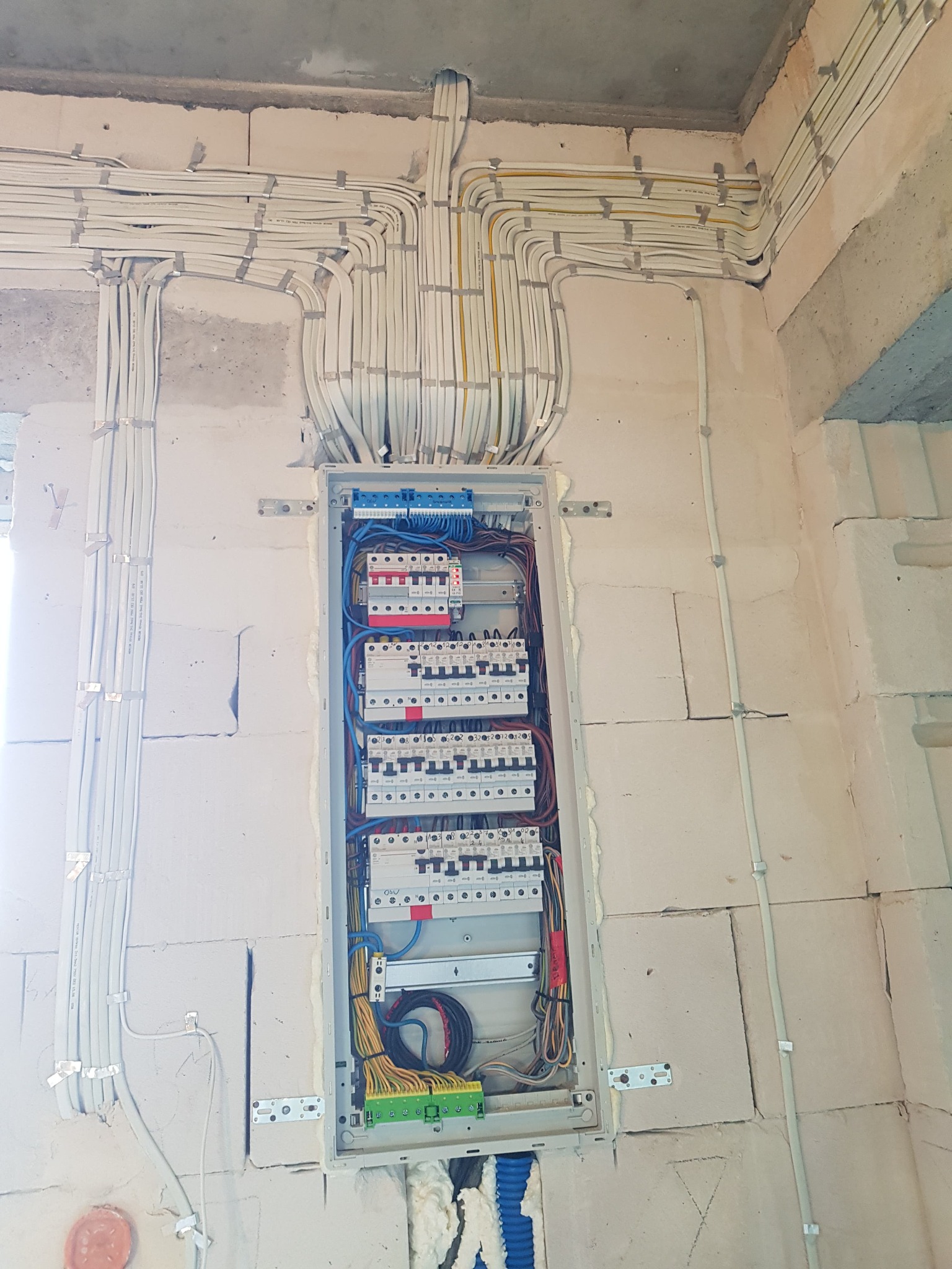 Rozdzielnica elektryczna z licznymi przewodami ułożonymi w korytach i przymocowanymi do surowej ściany z betonu komórkowego za pomocą metalowych uchwytów.