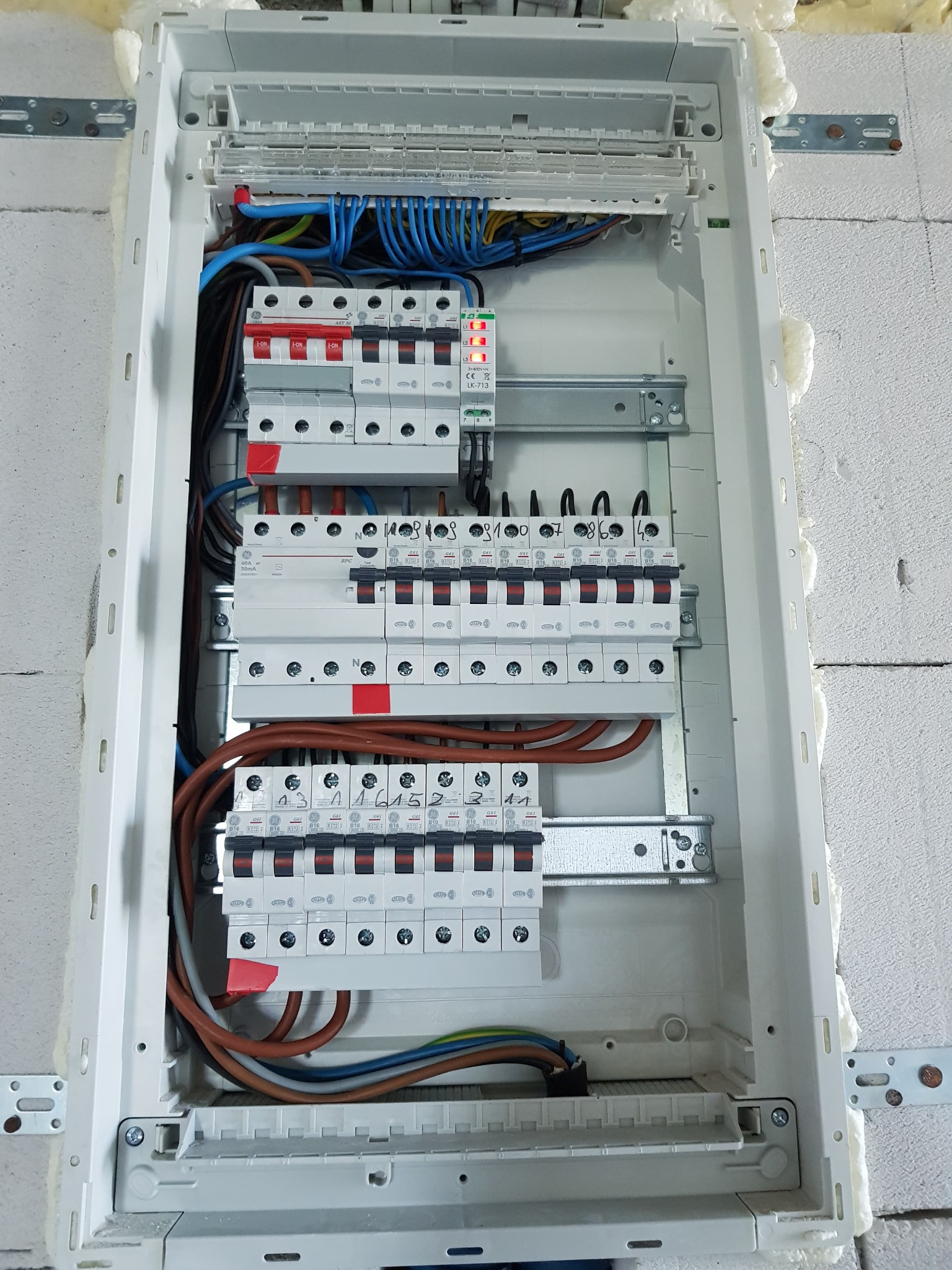 Otwarta skrzynka rozdzielcza z zamontowanymi bezpiecznikami i okablowaniem, widoczne ślady pianki montażowej na ścianie.