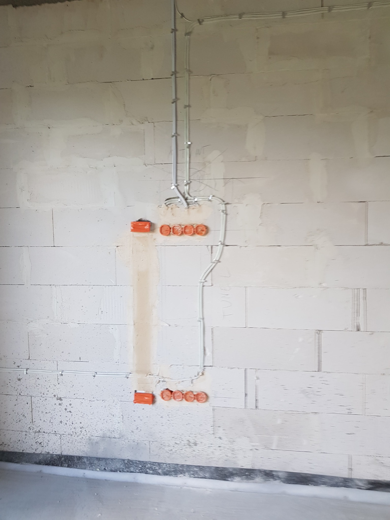 Świeżo położona instalacja elektryczna natynkowa z pomarańczowymi puszkami w surowym wnętrzu z białych bloczków, widoczne przewody przymocowane klamrami do ściany oraz ślady po tynkowaniu.