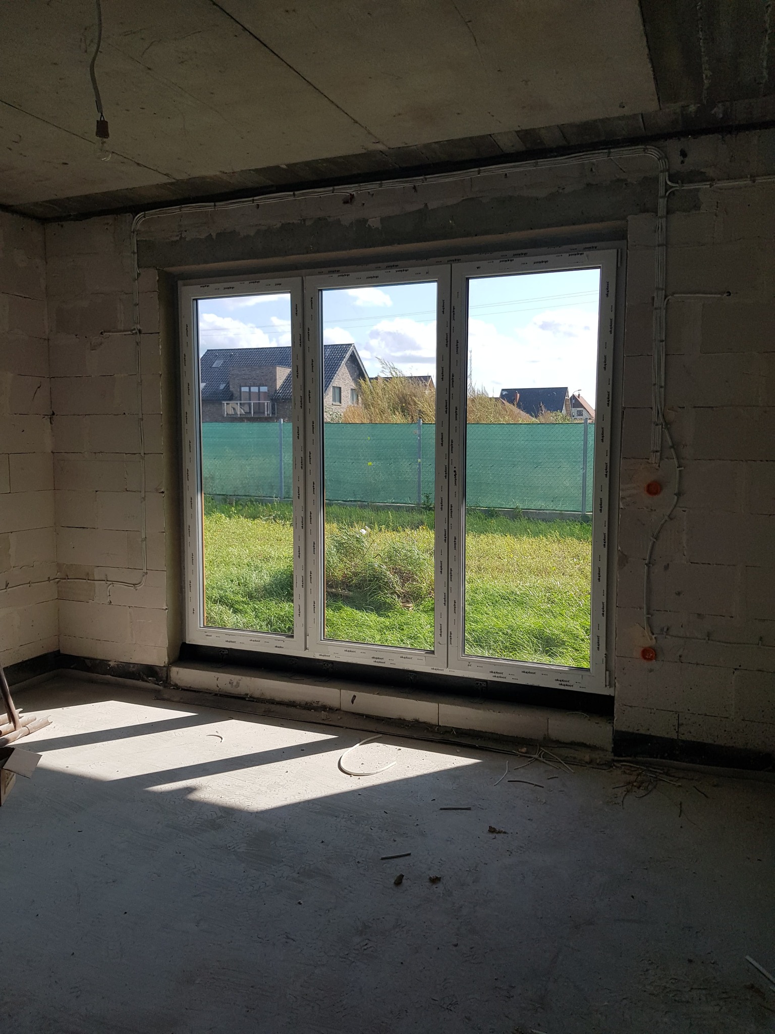 Surowy stan wnętrza z trzyskrzydłowym oknem balkonowym, widoczne przewody elektryczne na ścianie z betonu komórkowego i cień na podłodze.