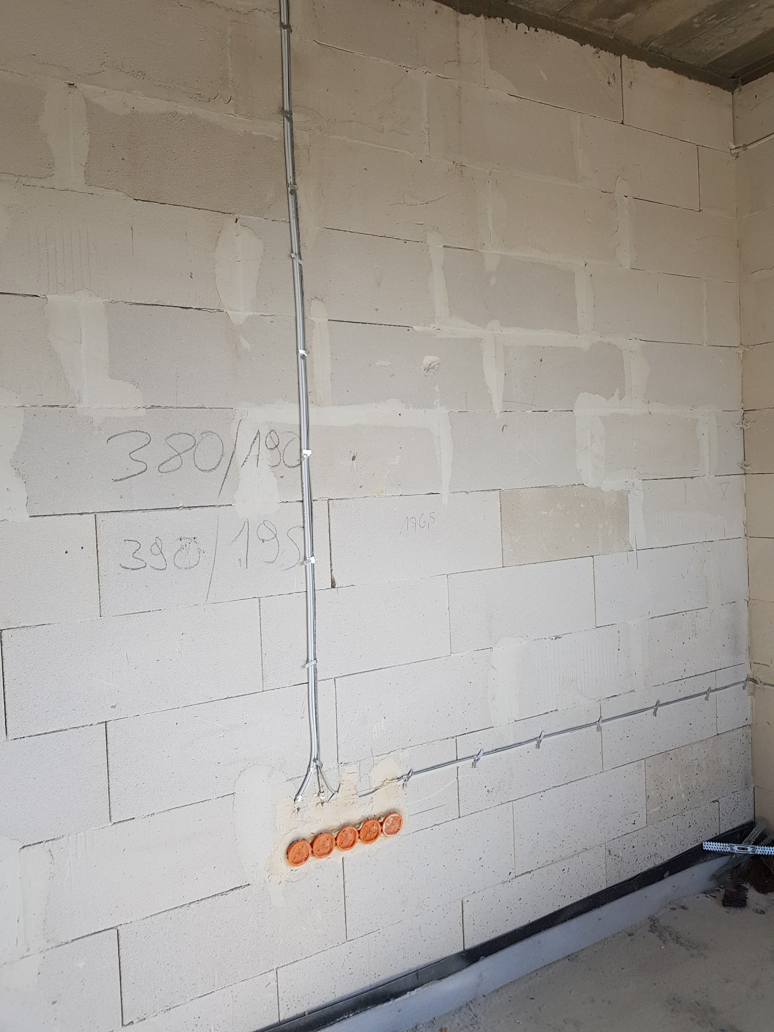 Surowa ściana z betonu komórkowego z instalacją elektryczną: kable w peszlu przymocowane uchwytami, puszka z pomarańczowymi otworami. Widoczne napisy ołówkiem na ścianie.