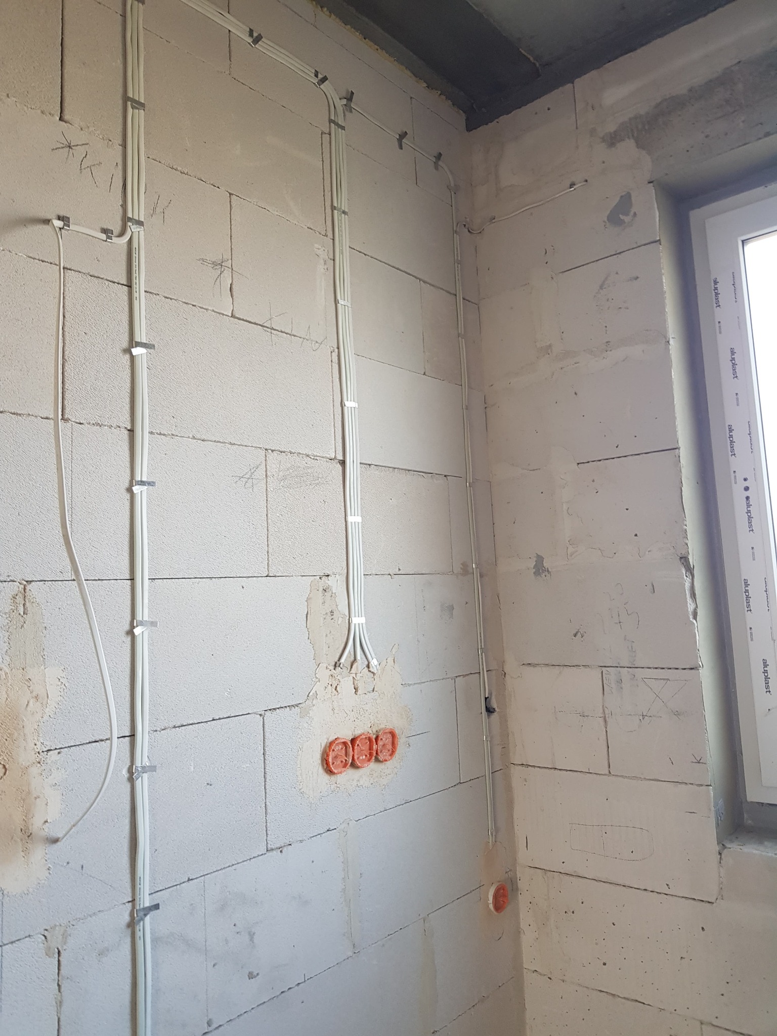 Białe przewody elektryczne ułożone pionowo na surowej ścianie z betonu komórkowego, zabezpieczone metalowymi uchwytami. Widoczne trzy pomarańczowe puszki instalacyjne podtynkowych gniazd...