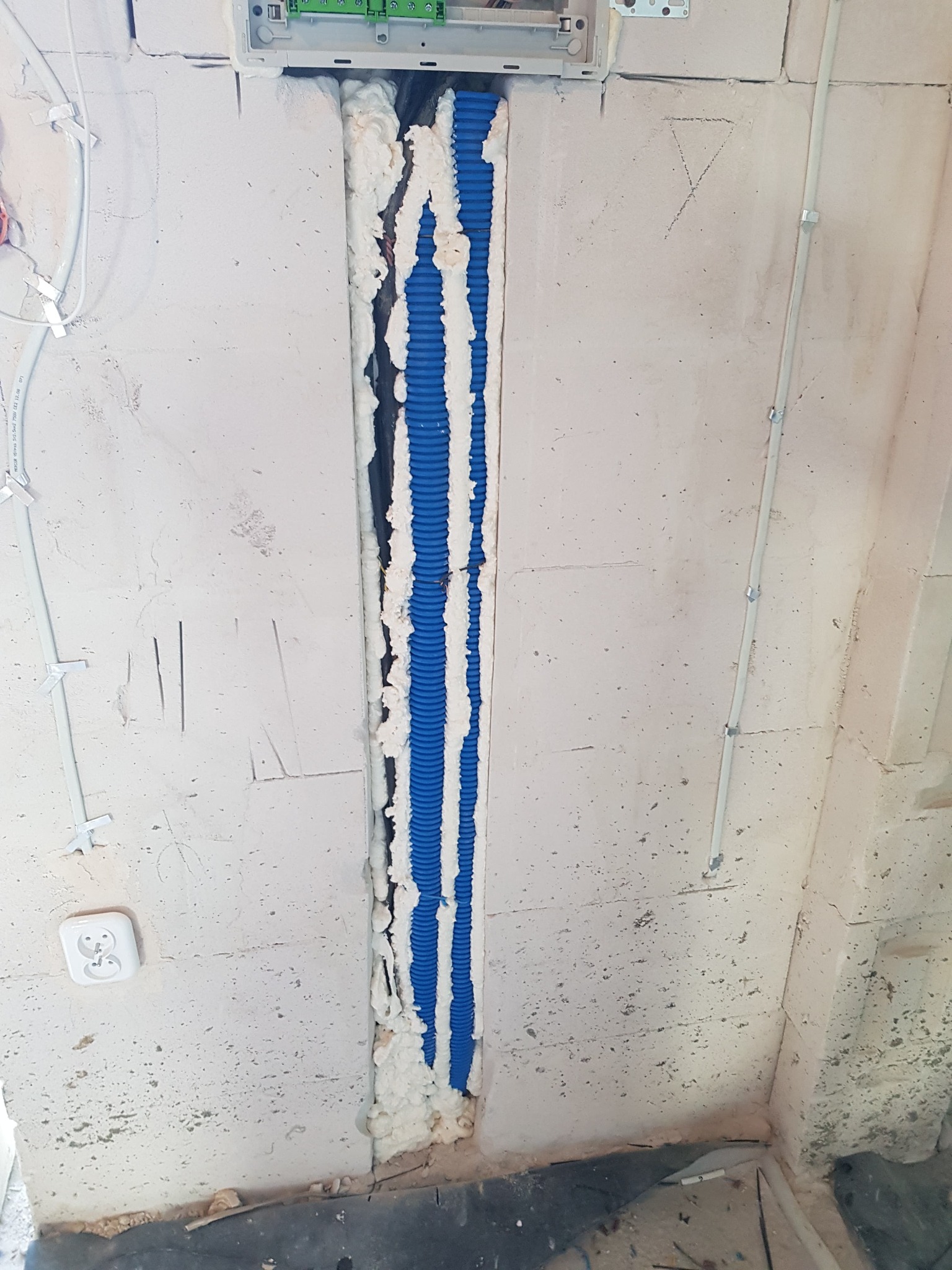 Wykuta bruzda w ścianie z betonu komórkowego, wypełniona niebieskimi rurami karbowanymi i pianką montażową, widoczne kable i gniazdko elektryczne w ścianie.