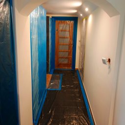 Usługi remontowo wykończeniowe - Korytarz zabezpieczony folią malarską przed remontem. Widoczne zakryte drzwi i podłoga. Odsłonięty włącznik elektryczny na ścianie. Przygotowania do malowania.