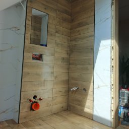 Usługi remontowo wykończeniowe - Łazienka w trakcie wykańczania: drewnopodobne płytki na ścianach i podłodze, białe płytki imitujące marmur, widoczne instalacje wodno-kanalizacyjne, narożnik pomieszczenia.