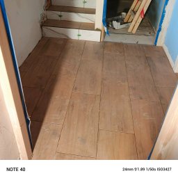 Usługi remontowo wykończeniowe - Ułożona podłoga z płytek imitujących drewno, widoczne schody z białych płytek z szarymi żyłami, zielone krzyżyki dystansowe, niebieska taśma malarska.