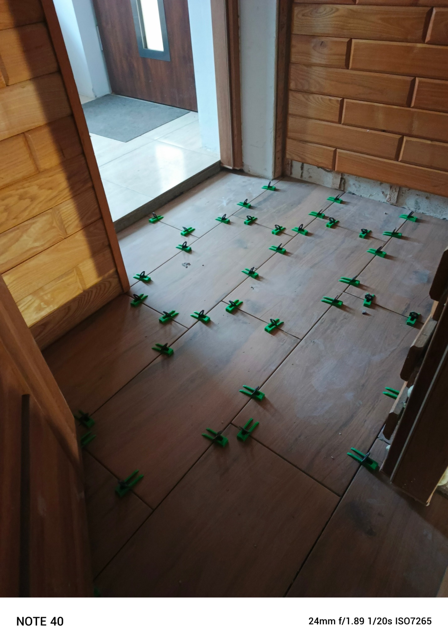 Układanie brązowych płytek podłogowych z użyciem zielonych klipsów poziomujących. Widoczne drewniane elementy wykończenia ścian i drzwi.