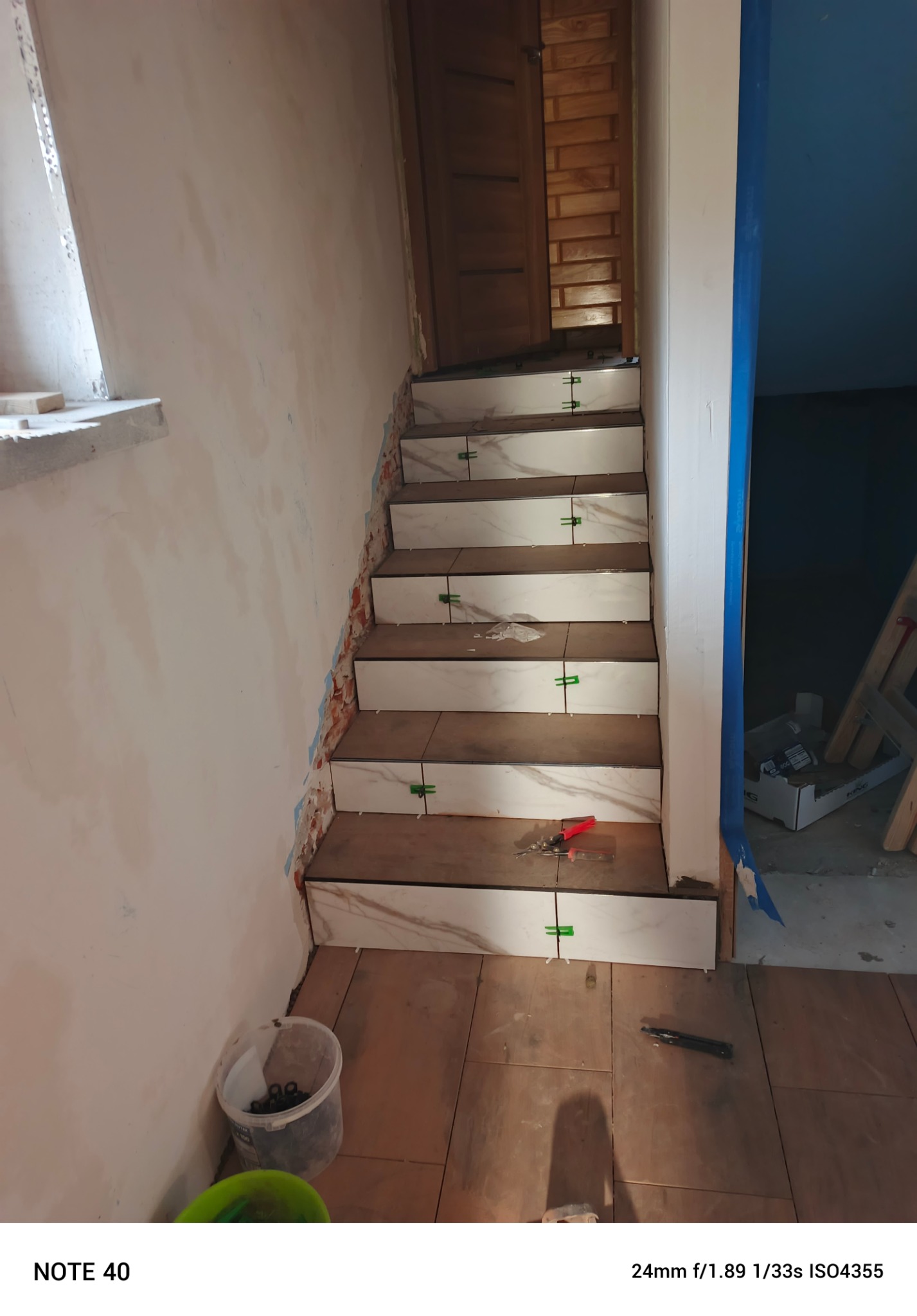 Układanie płytek na schodach z użyciem zielonych krzyżyków dystansowych. Widoczne narzędzia glazurnicze i wiadro z elementami montażowymi. Prace wykończeniowe w trakcie.