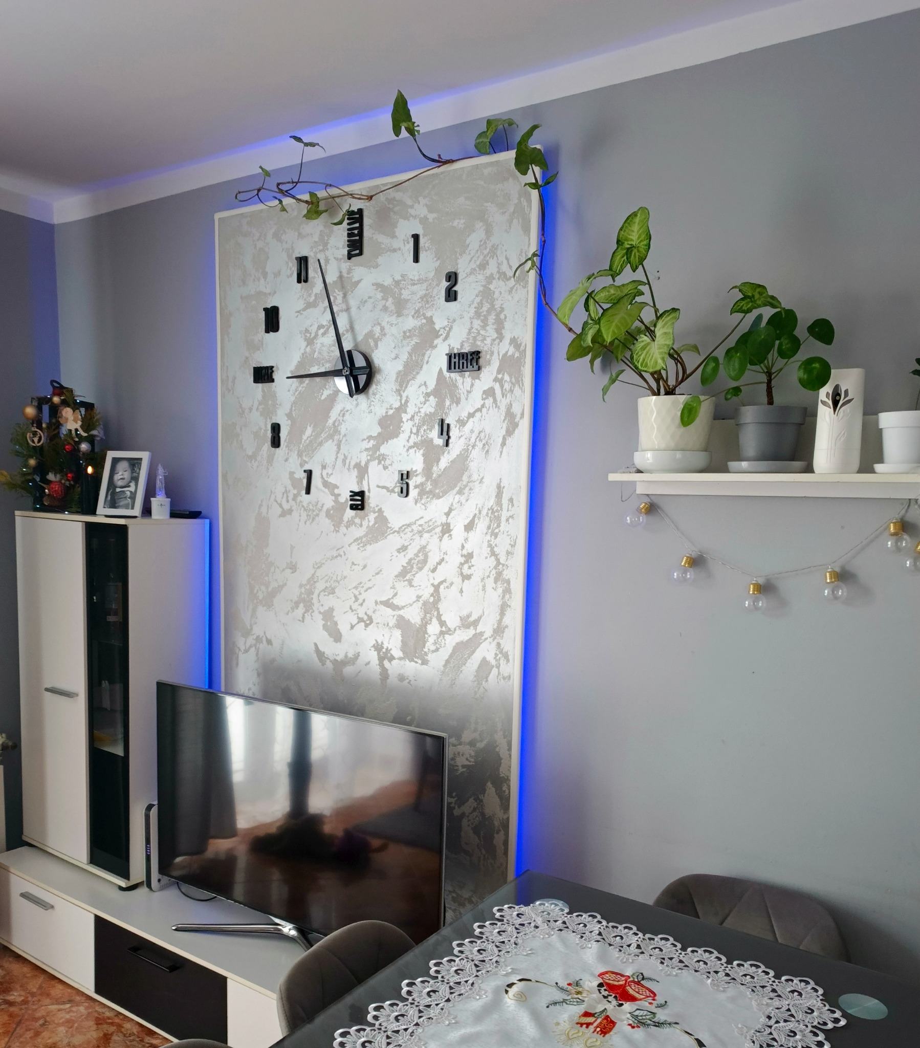 Szara ściana z dekoracyjnym panelem z efektem betonu, podświetlonym na niebiesko, z dużym zegarem. Półka z roślinami i komoda z telewizorem w salonie.