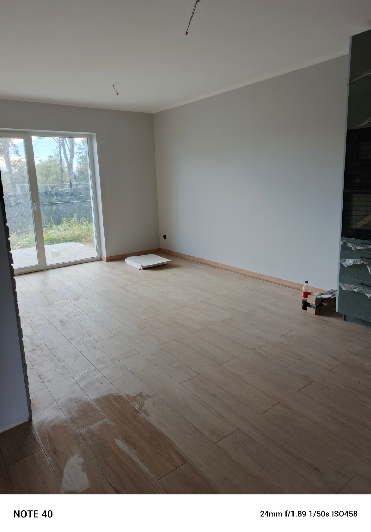 Jasne, wykończone wnętrze z drewnopodobną podłogą, szarą ścianą i dużym oknem balkonowym. Widoczne ślady po pracach wykończeniowych i resztki materiałów budowlanych.