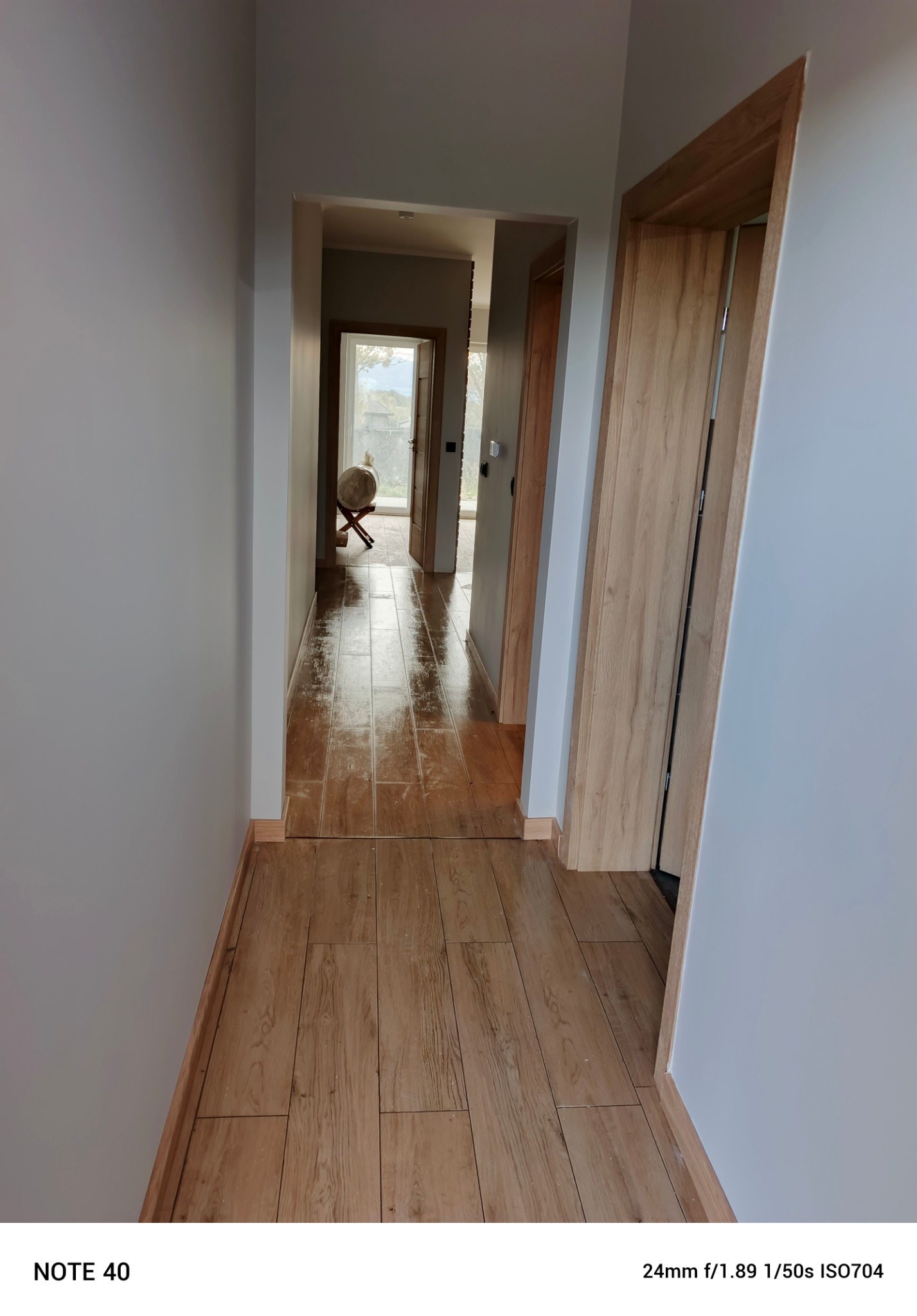 Korytarz po remoncie z jasnymi ścianami, drewnopodobną podłogą z płytek i nowymi drzwiami. Widoczne detale wykończeniowe, w tle otwarte drzwi do dalszych pomieszczeń.