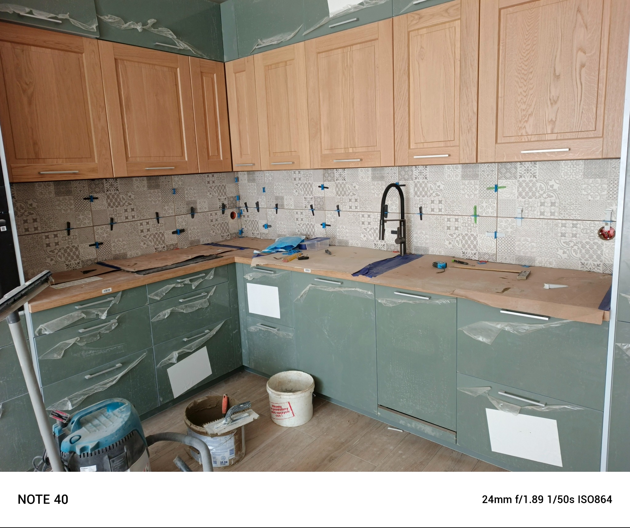 Montaż płytek ściennych w kuchni, widoczne krzyżyki dystansowe, zabezpieczone folią meble,  przygotowane narzędzia i materiały. Etap prac wykończeniowych.