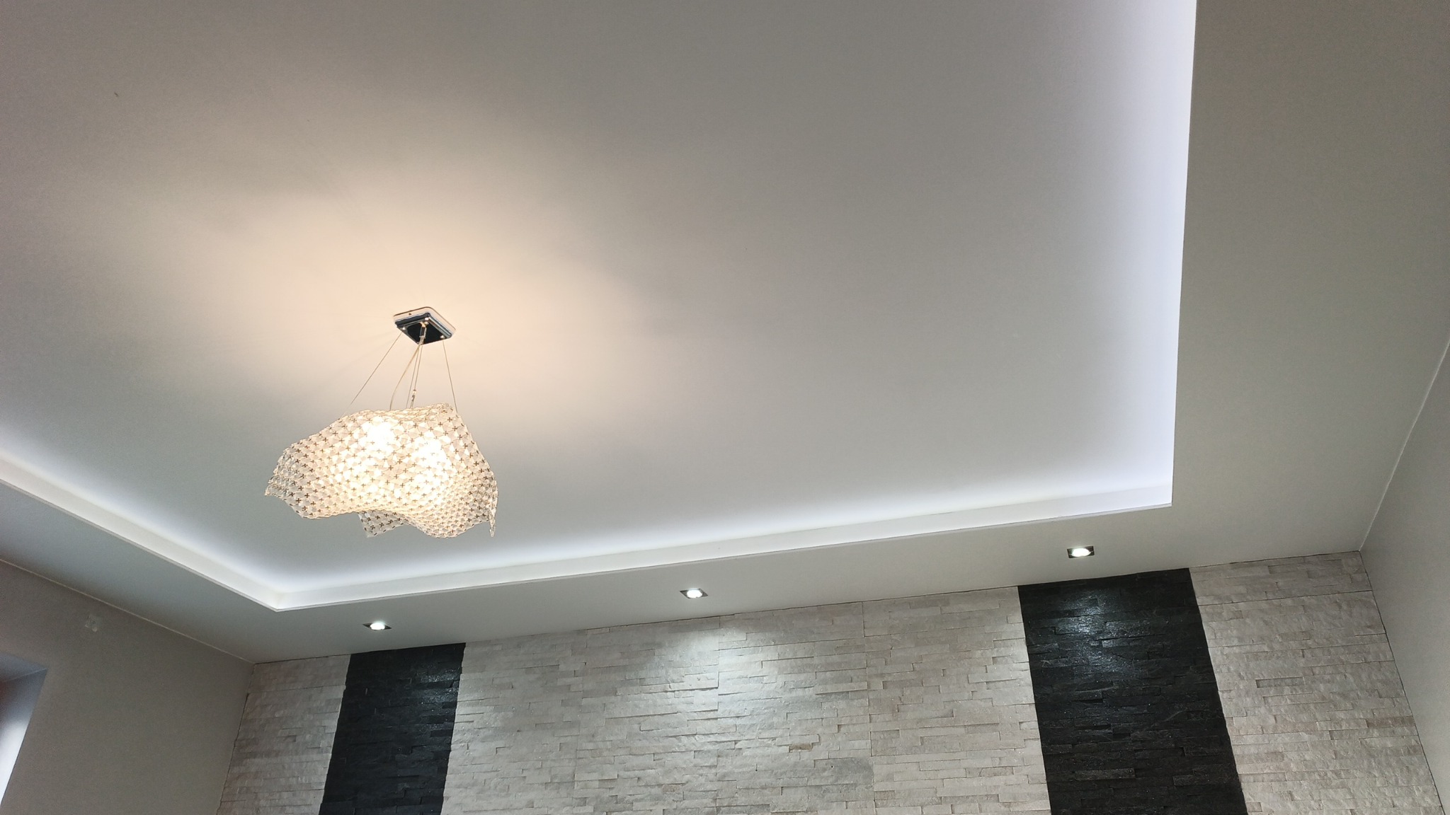 Nowoczesny sufit podwieszany z oświetleniem LED i dekoracyjnym żyrandolem. Ściana wykończona kamieniem dekoracyjnym z czarnymi akcentami. Minimalistyczny design.