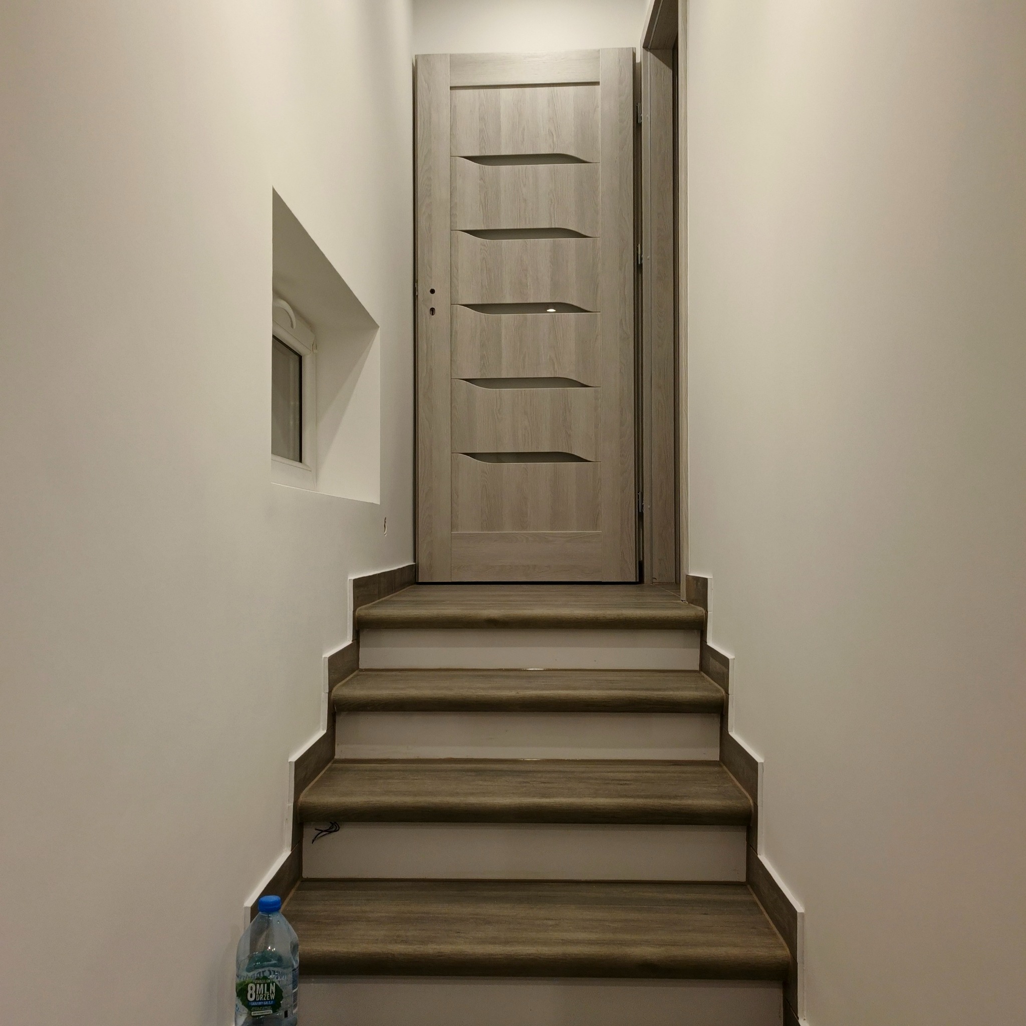 Schody z drewnopodobnymi panelami prowadzą do drzwi z poziomymi wstawkami. Białe ściany, małe okno. Butelka wody na dole schodów. Ujęcie od dołu.