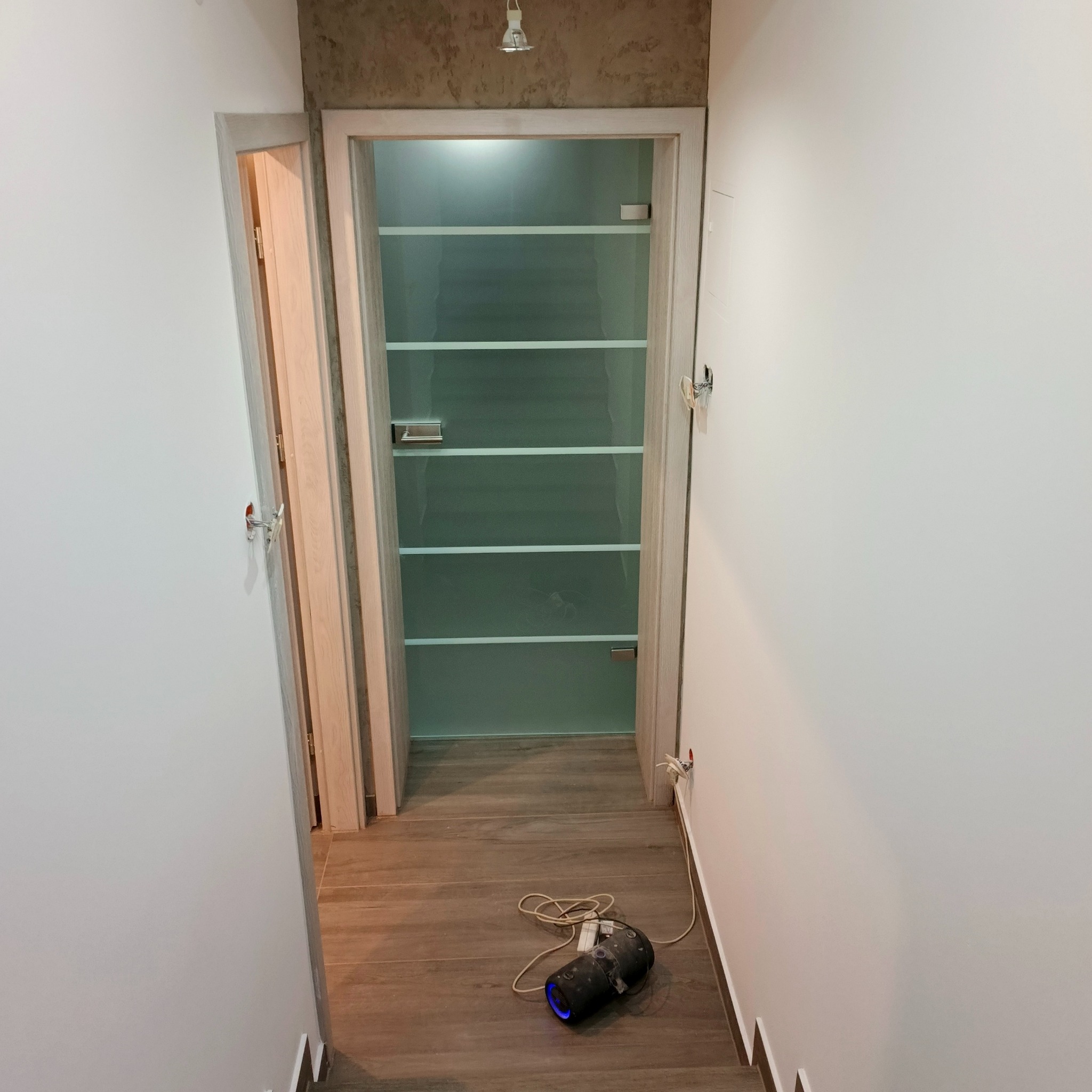 Korytarz po odświeżeniu z drewnopodobną podłogą i wnęką z półkami. Widoczne niedokończone gniazdko i lampa robocza na podłodze.