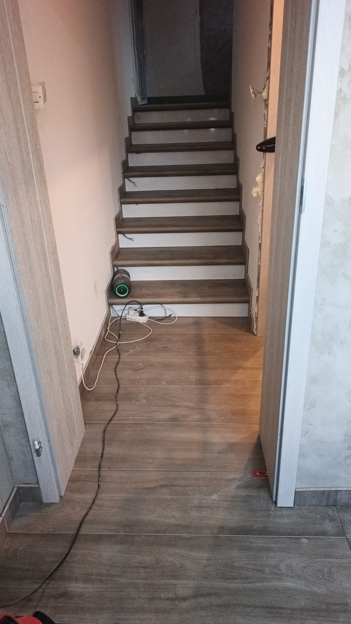 Wykończenie schodów z drewnopodobnymi panelami, widok z dołu. Ułożona podłoga z płytek. Widoczne narzędzia i kable. Częściowo wykończone ściany.