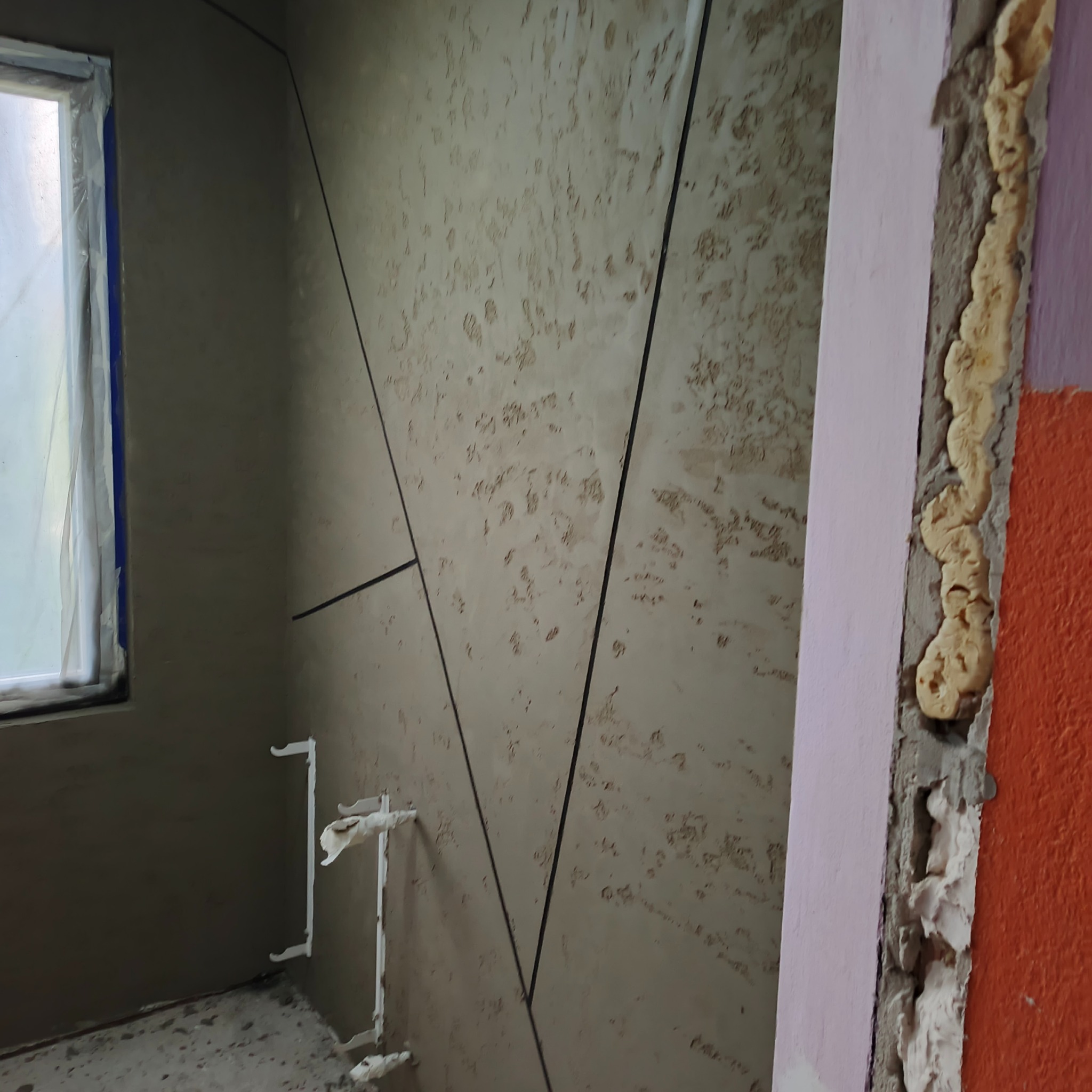 Ściana z dekoracyjną fakturą i czarnymi liniami, okno z folią ochronną, widoczne rury i pianka montażowa. Wnętrze w trakcie prac wykończeniowych.