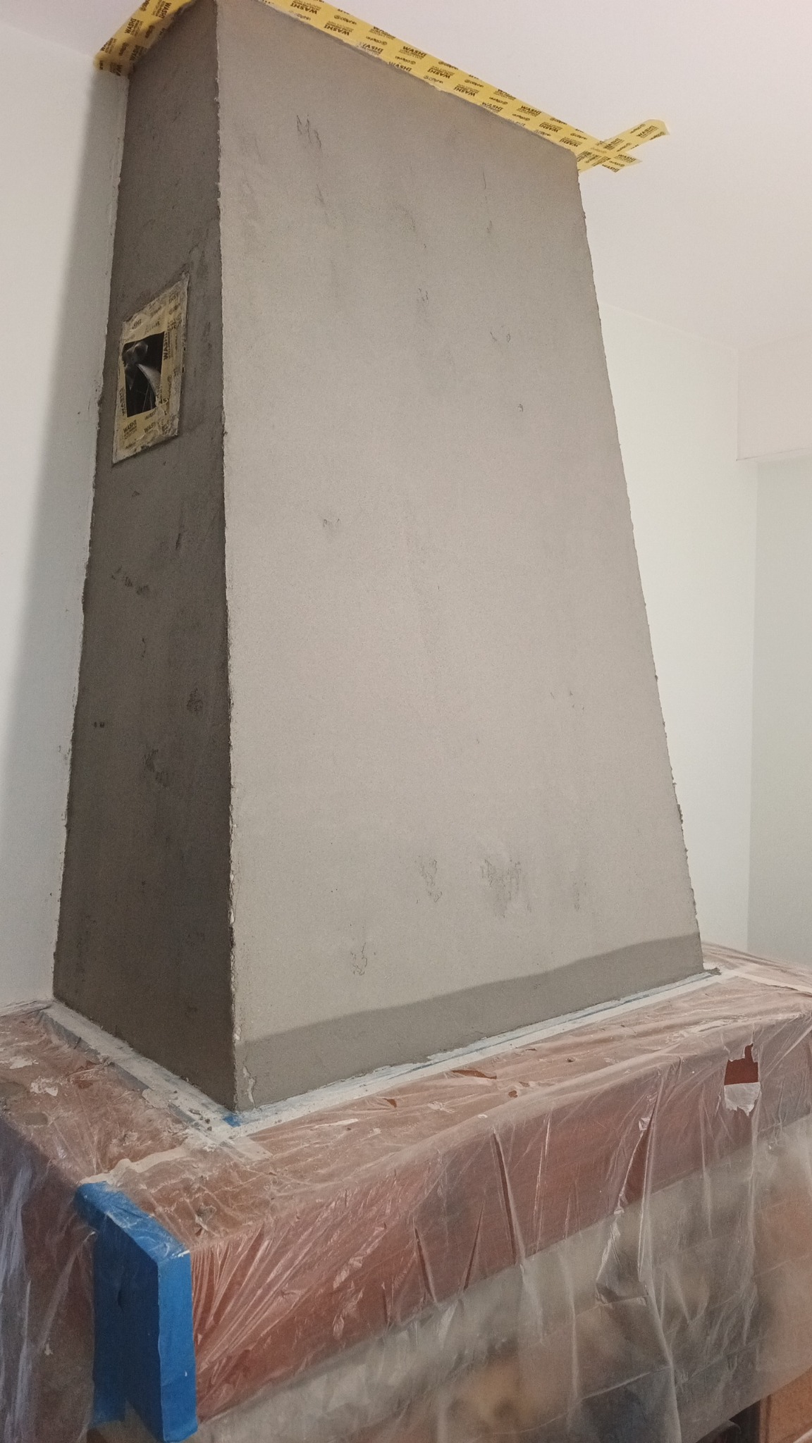 Narożny kominek w trakcie obkładania gładzią, zabezpieczony folią i taśmą malarską. Widoczny otwór rewizyjny. Ściany pomalowane na biało.