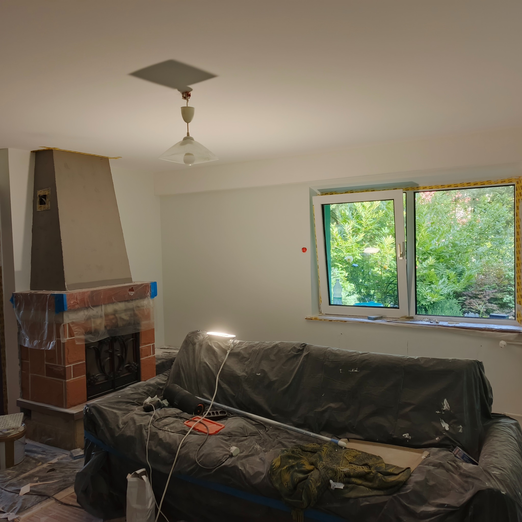 Pokój w trakcie odświeżania: zabezpieczony kominek, okno i meble folią malarską. Ściany przygotowane do malowania, widoczne taśmy malarskie.